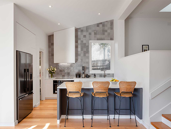 Modern kitchen with a back splash, bar stools at a white island, stainless steel appliances, and light wood flooring.
