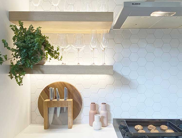 Kitchen with hexagonal tile backsplash, wooden shelves, glassware, knives, and oven with cookies baking.