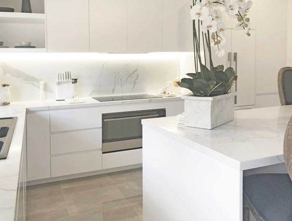 Modern white kitchen with marble countertops, white cabinets, a built-in stovetop, and an orchid plant on the island.