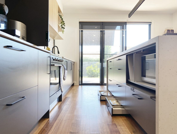 Modern kitchen with a long wooden floor, sleek cabinets, stove, and drawers, leading to a sunny windowed door.