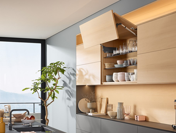 Modern kitchen with open wooden cabinets, glassware, and dishes. Large window with a view and a potted plant nearby.