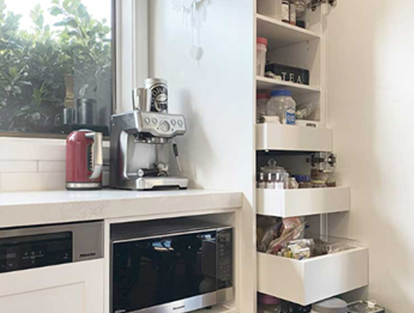 A kitchen corner with a coffee machine, kettle, microwave, and a pull-out pantry with jars and containers.