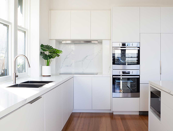 Minimalist white kitchen with wood floor, built-in ovens, and a potted plant by a window.
