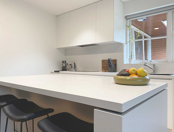 Minimalist kitchen with white cabinets, island, and stools. A fruit bowl sits on the island. Large window with light.