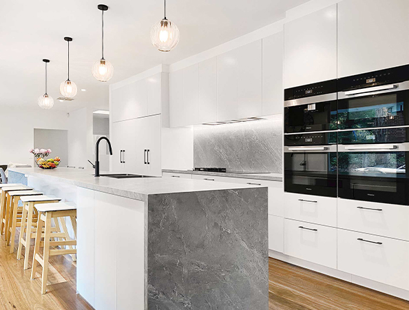 Modern kitchen with white cabinets, gray marble island, ovens, pendant lights, and wooden stools by a breakfast bar.