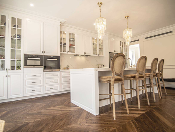 Spacious kitchen with white cabinets, a large island with bar stools, and elegant chandeliers on herringbone wood floors.