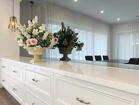 Elegant white kitchen counter with floral arrangement, mirrored backsplash, and pendant light. Sheer curtains in background.