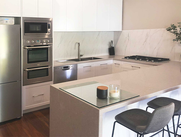 Modern kitchen with stainless steel appliances, marble backsplash, and two chairs at a light-colored island counter.
