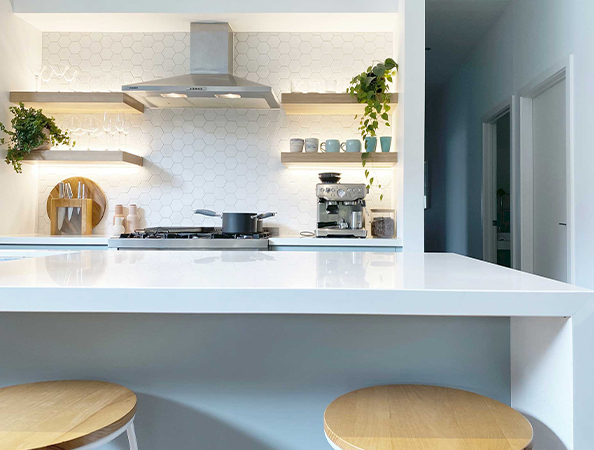 Modern kitchen with white countertops, hexagonal tile backsplash, wooden shelves, plants, and light wood stools. Modern kitchen with white countertops, hexagonal tile backsplash, wooden shelves, plants, and light wood stools.