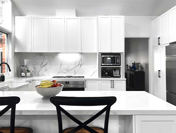 Modern white kitchen with marble backsplash, fruit bowl on counter, and black chairs. Appliances include oven and microwave. Modern white kitchen with marble backsplash, fruit bowl on counter, and black chairs. Appliances include oven and microwave.
