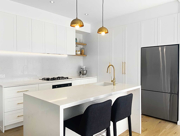 Modern kitchen with white cabinets, gold fixtures, island, two black chairs, and hanging pendant lights.