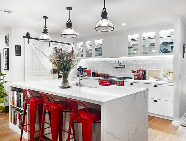 Modern kitchen with a white island, red stools, industrial lights, and glass cabinets displaying dishes.