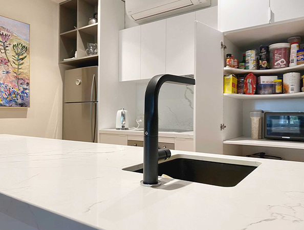 Modern kitchen with white cabinets, black faucet on island, open pantry with food items, and a colorful wall painting.