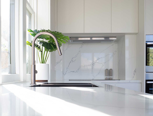 Modern kitchen with a sleek faucet, white countertops, marble backsplash, and a green plant by the window.