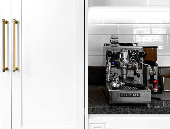 Espresso machine on a black countertop next to white cabinets with brass handles. Espresso machine on a black countertop next to white cabinets with brass handles.