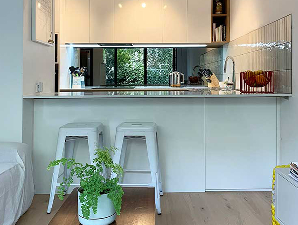 Compact modern kitchen with white cabinetry, two stools, a green plant, and a window overlooking greenery.