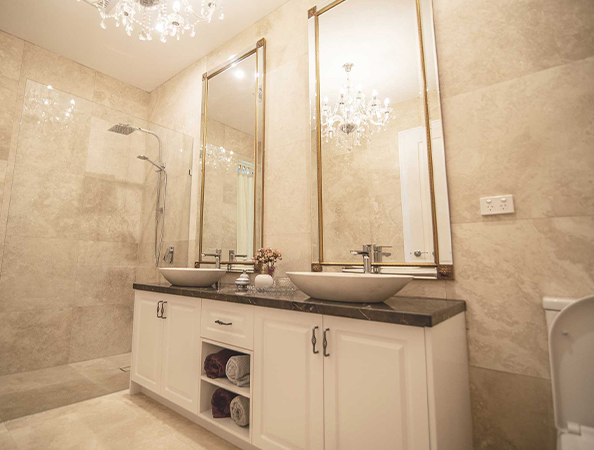 Elegant bathroom with double sinks, large mirrors, chandeliers, and a walk-in shower, featuring beige tiled walls. Elegant bathroom with double sinks, large mirrors, chandeliers, and a walk-in shower, featuring beige tiled walls.
