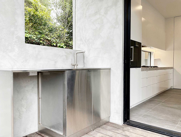 Minimalist kitchen with stainless steel counters, open glass window, and view of greenery outside.