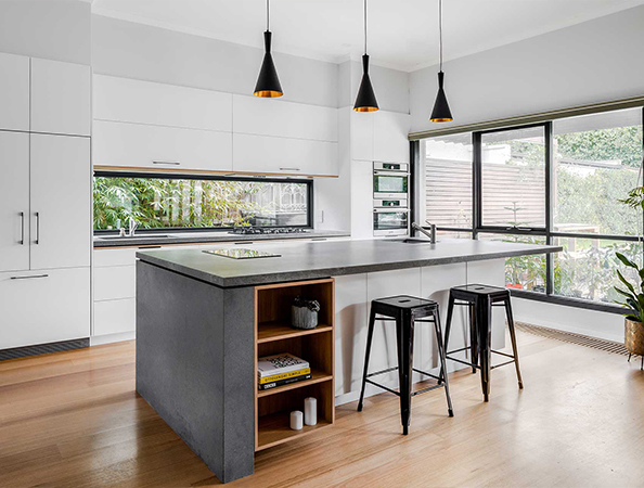 Modern kitchen with white cabinets, black pendant lights, a central island, and bar stools, overlooking a garden.