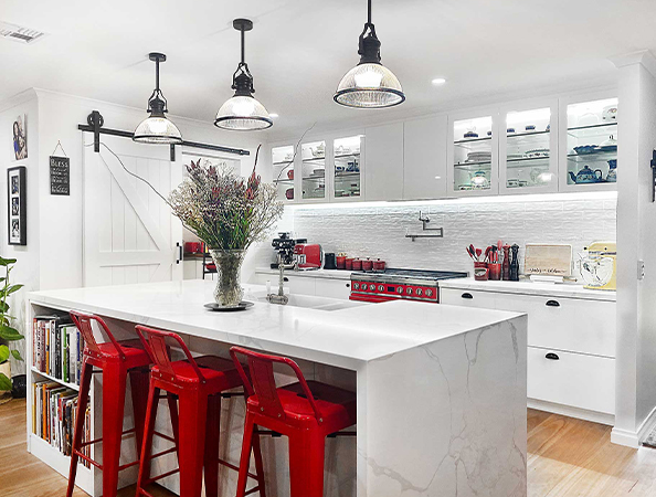 Modern kitchen with white cabinets, red stools, and a marble island. Hanging lights and a vase with flowers on the island.