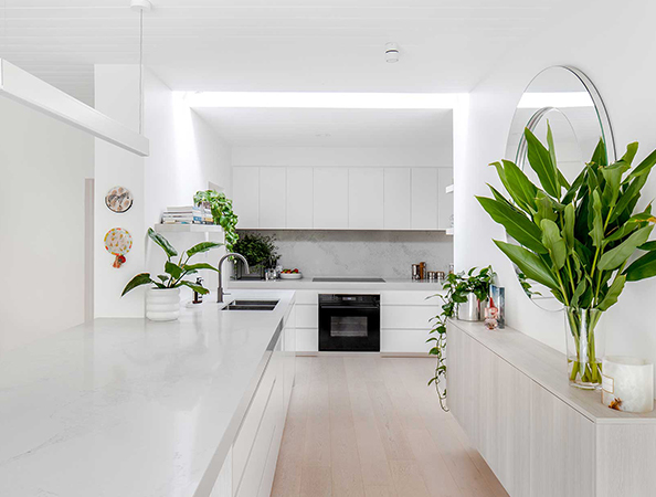 Bright, modern kitchen with white cabinetry, large plants, round mirror, and sleek countertops.