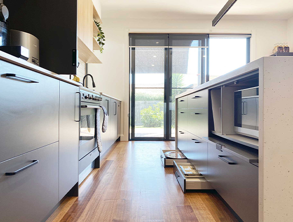 Modern kitchen with sleek dark cabinets, wood flooring, and sunlight streaming through a large glass door.
