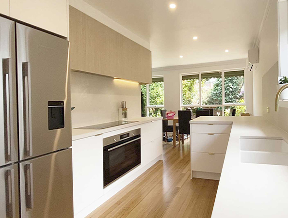 Modern kitchen with wooden flooring, white cabinets, stainless steel appliances, and large windows overlooking greenery.