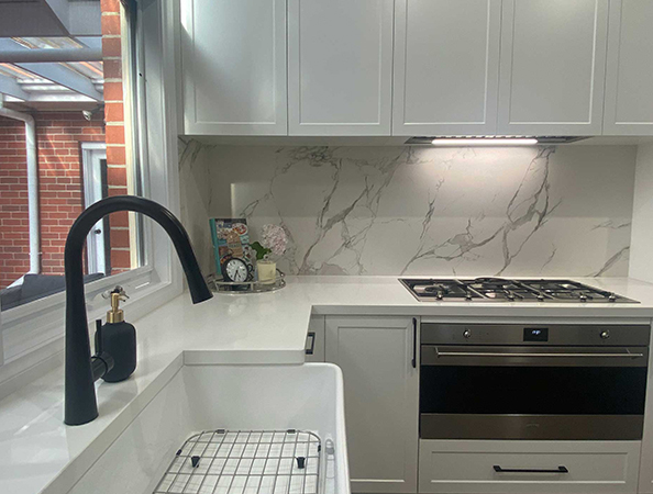 Modern kitchen with marble backsplash, black faucet, white countertops, and built-in gas stove under cabinets.