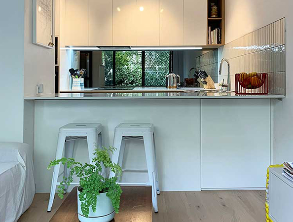 Modern kitchen with white cabinets, metal stools, and potted plant on wooden floor; large window in the background.