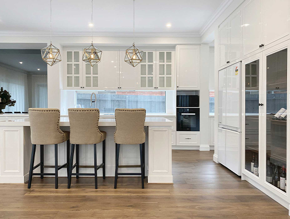 Modern kitchen with three beige barstools, white cabinets, pendant lights, and wood flooring.