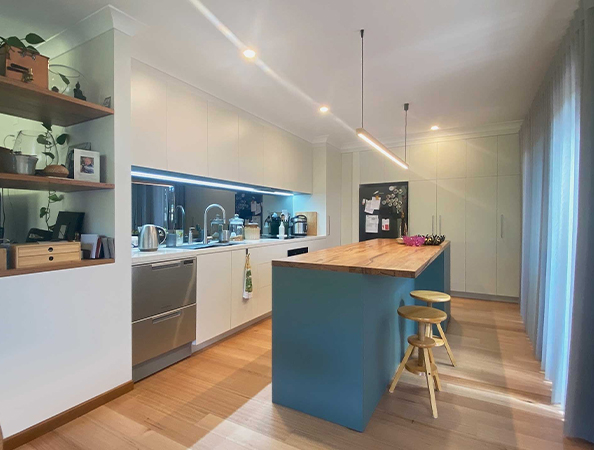 Modern kitchen with light wood floors, teal island with stools, white cabinetry, and plants on shelves.
