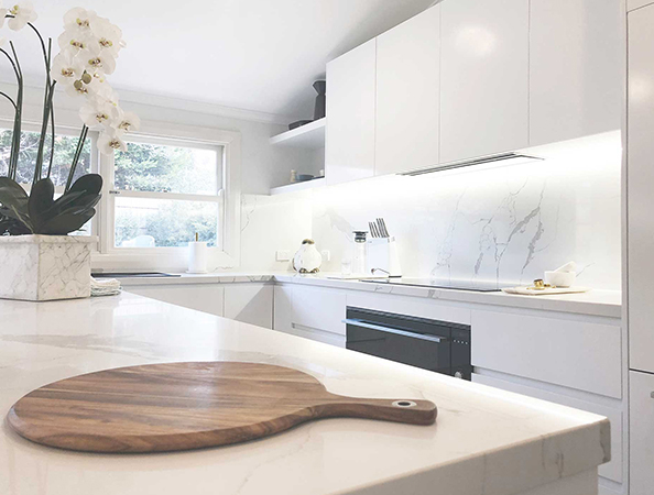 Minimalist white kitchen with marble countertops, orchids, wooden cutting board, and under-cabinet lighting.