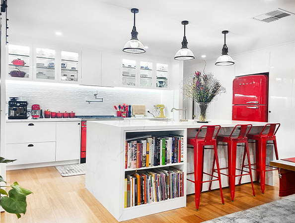 Modern kitchen with white cabinets, red fridge, red bar stools, and a white island with books and flowers.