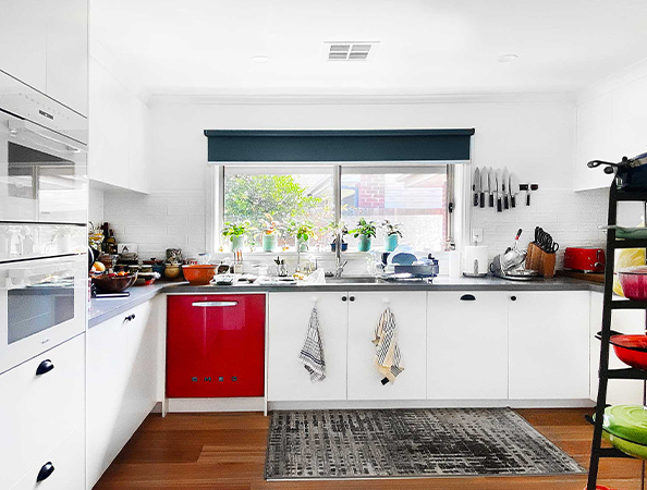 A bright, modern kitchen with white cabinets, a red dishwasher, and a window overlooking a garden.
