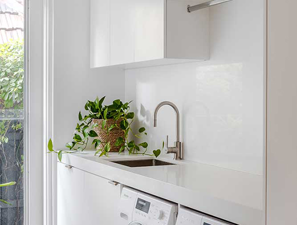 A modern laundry room with a white countertop, sink, washing machine, and a potted plant by the window.