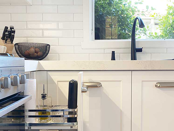 Modern kitchen with white cabinets, fruit bowl, spice rack, and black faucet near a window with greenery outside.