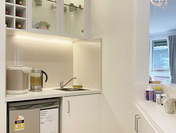 Compact kitchen with white cabinets, a small sink, and a mini-fridge. Upper shelves hold glasses and a wine rack.