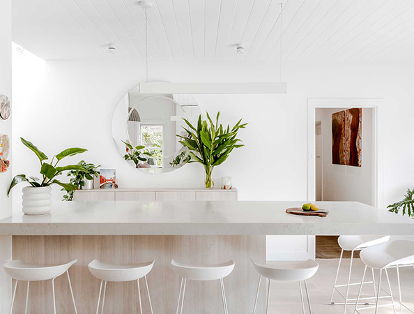 Minimalist white kitchen with a large mirror, green plants, and modern white barstools around a marble island.