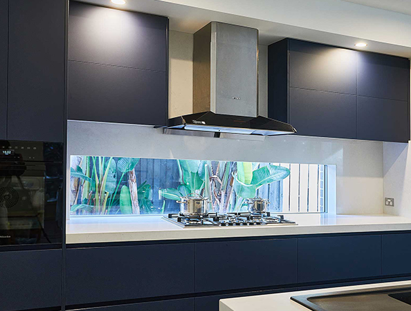 Modern kitchen with dark cabinets, stainless steel stove and hood, and a window view of green plants outside.