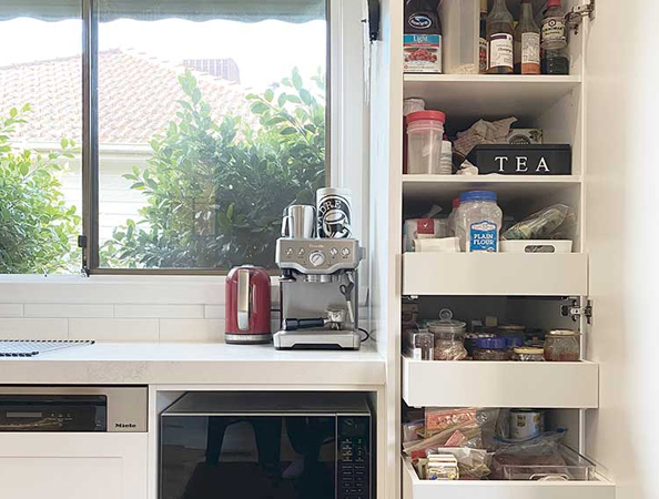 Kitchen with coffee machine, kettle, and open pantry shelves next to a window with bushes outside. Kitchen with coffee machine, kettle, and open pantry shelves next to a window with bushes outside.