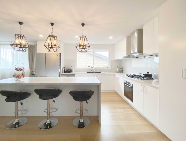 Modern white kitchen with a large island, three stools, pendant lights, and stainless steel appliances.