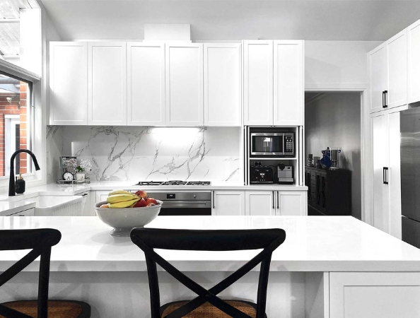 Modern white kitchen with marble backsplash, island, fruit bowl, and black chairs. Stainless appliances visible.
