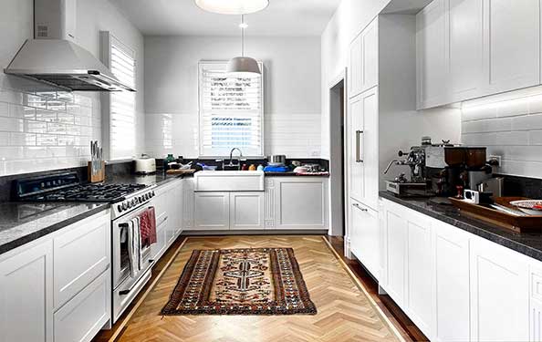 Bright kitchen with white cabinets, black countertops, a farmhouse sink, herringbone wooden floor, and a patterned rug.