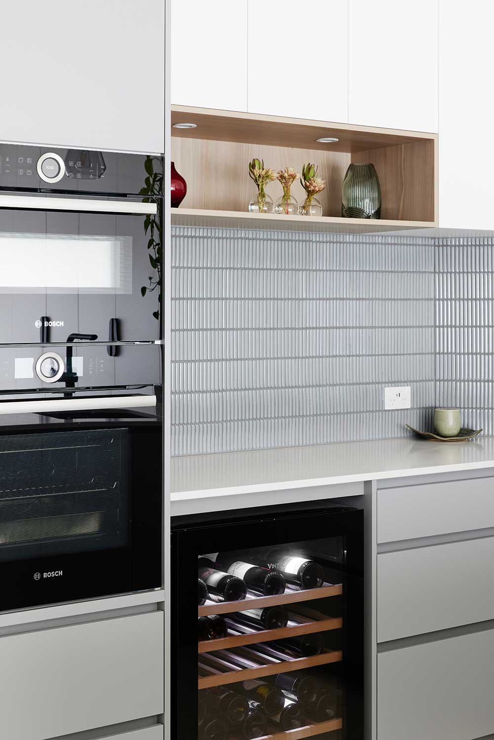 Modern kitchen with built-in ovens, wine cooler, gray tile backsplash, and decorative shelves holding vases and plants. Modern kitchen with built-in ovens, wine cooler, gray tile backsplash, and decorative shelves holding vases and plants.
