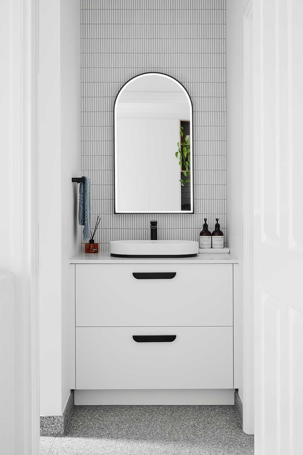 Minimalist bathroom with a white vanity, oval mirror, and decorative items on the countertop against a tiled wall. Minimalist bathroom with a white vanity, oval mirror, and decorative items on the countertop against a tiled wall.