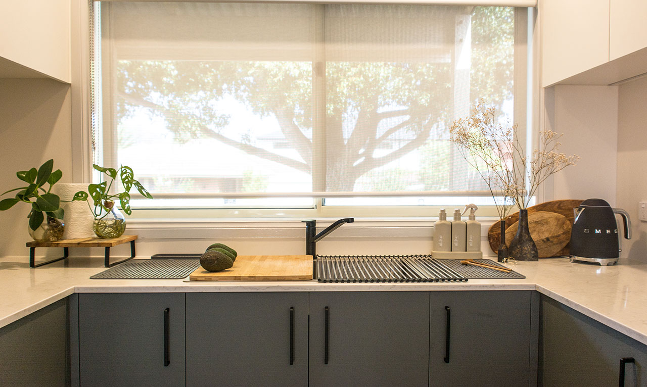 Modern kitchen with plants, cutting board, and avocado on countertop. Large window with tree view in background. Modern kitchen with plants, cutting board, and avocado on countertop. Large window with tree view in background.