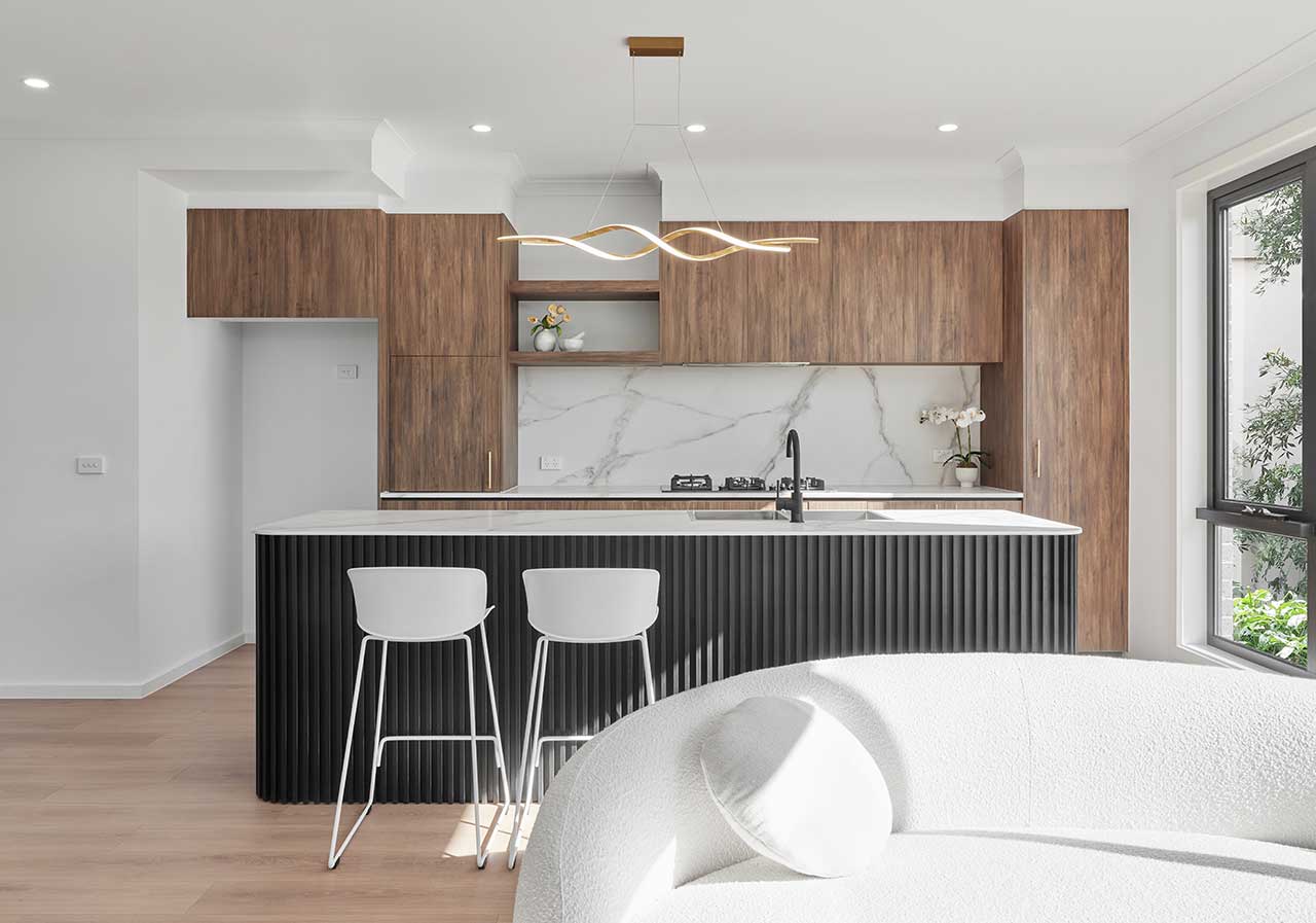 Modern kitchen with wood cabinets, marble backsplash, black island, white stools, and curved white sofa.