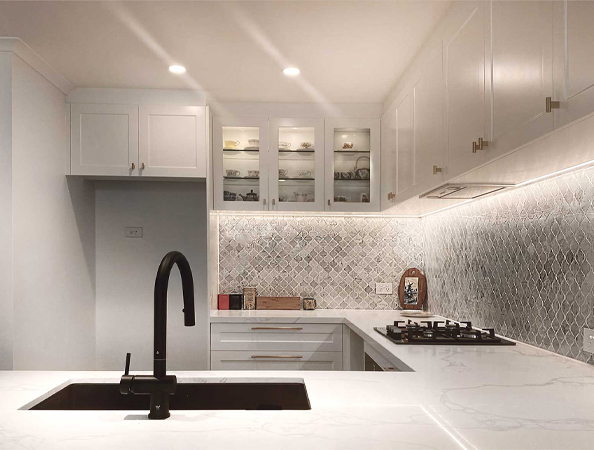 Modern kitchen with a black faucet, white cabinets, marble countertops, and tiled backsplash under ambient lighting.