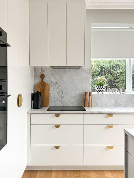 Modern kitchen with white cabinets, marble backsplash, and wooden accents. Bright window view with greenery outside. PROJECT LUX AND LIGHT @ BRIGHTON EAST