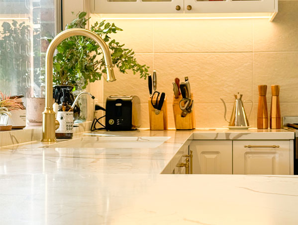 Modern kitchen with marble countertops, gold faucet, knife block, toaster, and plants on the windowsill.